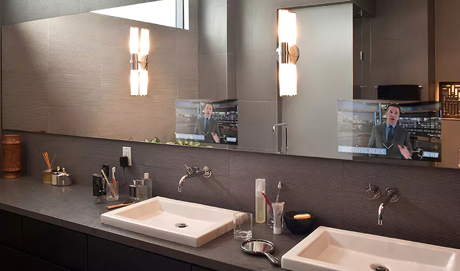 Full-length mirror TV on a wall displaying newscaster above two white basins.